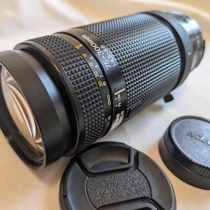 Könnte beinhalten: Ein schwarzes Nikon 75-300mm Kameraobjektiv mit zwei Objektivdeckeln. Das Objektiv hat einen strukturierten Griff und Markierungen für Brennweite und Blende. Die Objektivdeckel sind schwarz und rund, mit dem Nikon-Logo.