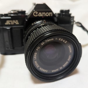 以下が含まれることがあります： 黒いキヤノンAV-1フィルムカメラ、35-70mmレンズ付き。レンズには「Canon Lens FD Zoom 35-70mm 1:3.5-4.5 Made in Japan」と書かれています。カメラには黒いストラップが付いています。カメラは白い面にあります。