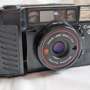 以下が含まれることがあります： 38mmレンズを搭載した黒いキヤノンクォーツデート35mmフィルムカメラ。カメラには内蔵フラッシュがあり、レンズには「Canon Lens 38mm 1:2.8 Auto Focus」の文字が印刷されています。白い表面にあります。