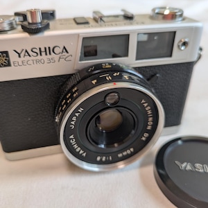 Puede incluir: Una cámara vintage Yashica Electro 35 FC con cuerpo negro y plateado y una lente Yashinon DX 40mm f/2.8. La cámara tiene el texto "YASHICA ELECTRO 35 FC" impreso en la parte delantera. También hay una tapa de lente negra con el logotipo de Yashica.