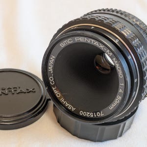 May include: A black SMC Pentax-M Macro 50mm f/4 camera lens with a matching lens cap. The lens has a ribbed focus ring and markings. The lens cap is round with the Pentax logo.