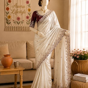 May include: A white satin saree featuring a deep maroon blouse with intricate silver embroidery along the neckline and sleeves. The saree has a matching maroon border with detailed silver floral patterns and small scattered embellishments.