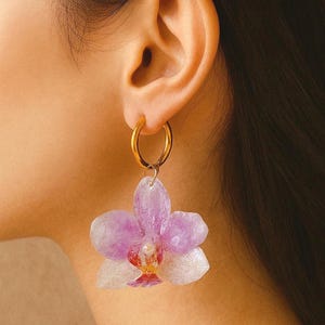 以下が含まれることがあります： ゴールドのフープピアス。薄紫と白の蘭の花のペンダントが付いています。花の中心部は赤と黄色のアクセントで詳細に表現され、自然でエレガントな外観を演出しています。スタイリッシュなアクセサリーです。