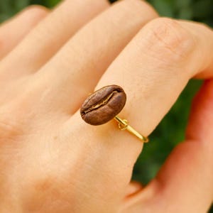 May include: A gold ring with a coffee bean as its focal point. The coffee bean is a deep brown hue, displaying a central crease. The ring is worn on a finger, set against a blurred green backdrop.