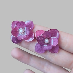 Könnte beinhalten: Ein Paar Ohrstecker mit Blumenmuster. Jeder Ohrring zeigt eine tiefviolette Hortensienblüte mit einem Perlenmittelpunkt. Die Blütenblätter haben eine zarte, geaderte Textur und sind in klarem Harz eingeschlossen.