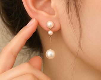 May include: Elegant gold drop earrings featuring three white pearls. The top two pearls are close to the ear, with a larger pearl suspended below on a delicate gold chain. The earrings are a classic jewelry piece.