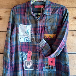 May include: A long-sleeved, button-down flannel shirt in a plaid pattern of blue, purple, and brown. The shirt features several patches with various designs, including a cat portrait, floral patterns, and a red star. The cuffs and collar are also patched.