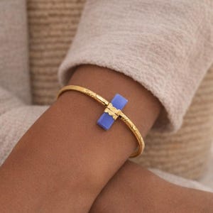 Peut inclure: Bracelet jonc doré avec une bande texturée et une pièce maîtresse rectangulaire bleu clair. Le bracelet est porté sur un bras, avec une manche de couleur neutre en arrière-plan. Le bijou est un accessoire élégant.