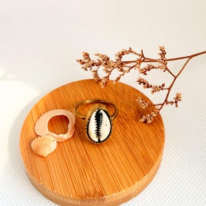 May include: A gold-coloured ring with a cowrie shell centre, displaying black and white patterns. The ring is presented on a round wooden coaster, alongside small seashells and dried flowers. The design suggests a coastal or bohemian style.