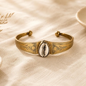 Può includere: Un bracciale a polsino color oro con un motivo geometrico inciso e una conchiglia di ciprea centrale. Il bracciale ha estremità arrotondate e un design decorativo. La conchiglia è bianca con segni marroni. Il bracciale è esposto su un tessuto color neutro.