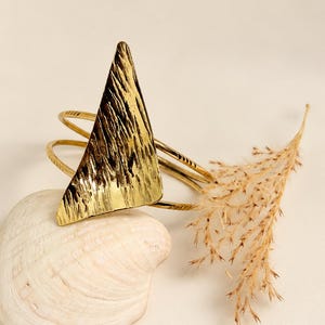 Op de afbeelding: Een goudkleurige metalen armband met een groot, driehoekig middendeel met een getextureerd, gehamerd oppervlak. De band bestaat uit twee dunne, parallelle draden met kleine, gegraveerde markeringen langs de zijkanten van de structuur.