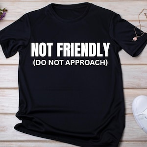Op de afbeelding: Zwart T-shirt met de witte tekst "NOT FRIENDLY (DO NOT APPROACH)". Het shirt heeft een ronde hals en korte mouwen. De stof lijkt een zacht, lichtgewicht materiaal te zijn.