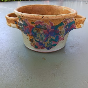 May include: A handmade ceramic bowl with two side handles. The bowl features a speckled tan rim and exterior, with colorful abstract paint splatters in shades of blue, pink, and green on a cream-colored base. The bowl is suitable for serving or display.