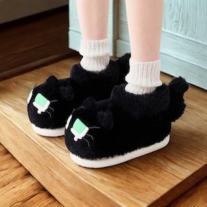 Puede incluir: Zapatillas negras con forma de gato, suelas blancas y detalles de ojos verdes. Las zapatillas tienen una textura suave y un diseño de cola. La persona lleva calcetines blancos. Las zapatillas están sobre una superficie de madera.