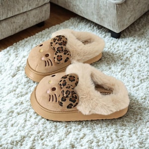 Op de afbeelding: Een paar bruinbeige pantoffels met een cartoon kattengezicht ontwerp. De pantoffels hebben een pluizige, lichtbruine rand en een luipaardprint strik. De pantoffels liggen op een wit tapijt.