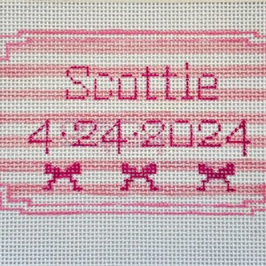 May include: A hand-stitched canvas with the name "Scottie" and the date "4-24-2024" embroidered in pink. The design includes a pink frame, horizontal stripes, and three small bow accents.