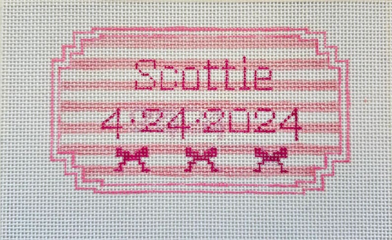 May include: A hand-stitched canvas with the name "Scottie" and the date "4-24-2024" embroidered in pink. The design includes a pink frame, horizontal stripes, and three small bow accents.