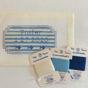 May include: A needlepoint canvas with the name "Pusher" and the date "1-20-2020" stitched in blue. Three spools of Tilli Tomas Essentials yarn in white, light blue, and dark blue are displayed next to the canvas.