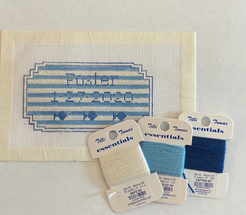 May include: A needlepoint canvas with the name "Pusher" and the date "1-20-2020" stitched in blue. Three spools of Tilli Tomas Essentials yarn in white, light blue, and dark blue are displayed next to the canvas.