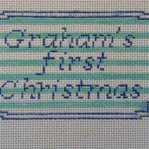 May include: Needlepoint canvas with the text "Graham's First Christmas" in dark blue stitching. The text is inside a light blue and white striped rectangle with a dark blue border. The background is a white canvas.