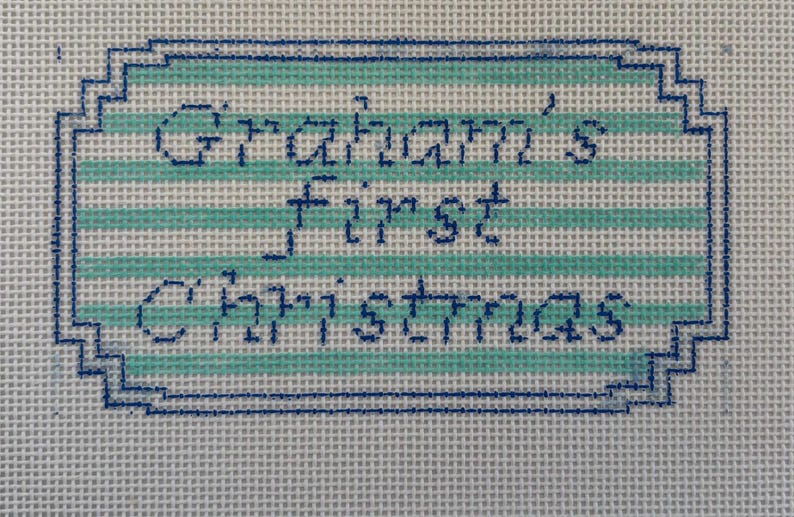 May include: Needlepoint canvas with the text "Graham's First Christmas" in dark blue stitching. The text is inside a light blue and white striped rectangle with a dark blue border. The background is a white canvas.