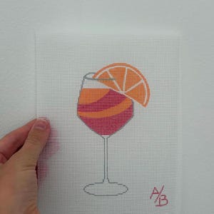 Könnte beinhalten: Eine Stickleinwand mit einem Motiv eines Weinglases, gefüllt mit einem geschichteten orangefarbenen und rosafarbenen Cocktail, garniert mit einer Orangenscheibe. Die Initialen A/B sind unten rechts in Rot aufgestickt.
