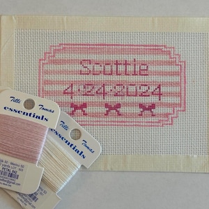 May include: A cross-stitch project with the name "Scottie" and the date "4-24-2024" in pink thread. Three spools of embroidery floss in shades of pink and white are in the foreground.