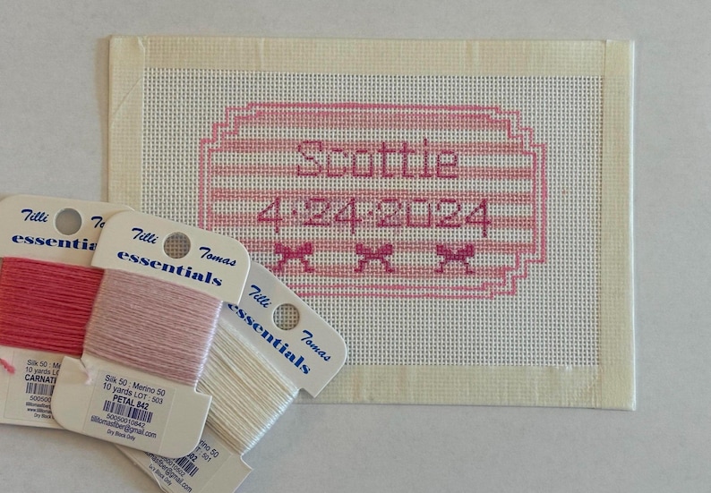 May include: A cross-stitch project with the name "Scottie" and the date "4-24-2024" in pink thread. Three spools of embroidery floss in shades of pink and white are in the foreground.