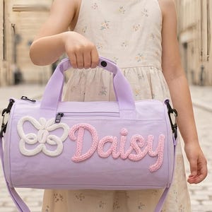 Puede incluir: Una bolsa de deporte lavanda con un diseño de mariposa blanca y el nombre "Daisy" en letras rosas. La bolsa tiene una asa superior y una correa para el hombro. La bolsa es sostenida por una persona.