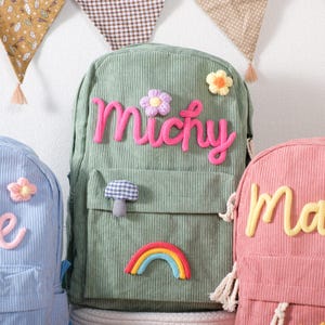 May include: A green corduroy backpack with the name "Michy" in pink yarn, decorated with a flower, mushroom, and rainbow. Other backpacks in blue and pink are also visible, each with a name and decorative elements.