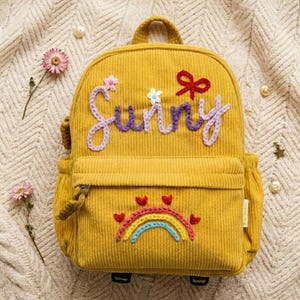 May include: Mustard yellow corduroy backpack with the word "Sunny" embroidered in purple. The backpack features a rainbow and heart design, a top handle, and side pockets. A red bow and small flowers are also embroidered on the front.