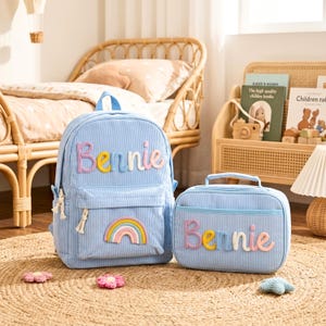 May include: A light blue corduroy backpack and a matching lunch bag, both featuring the name Bennie in colorful, raised, textured lettering. The backpack includes a rainbow patch on the front pocket, and both items have white rope zipper pulls.