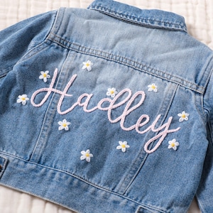May include: A light blue denim jacket with the name "Hadley" embroidered in pink script on the back. Small white and yellow daisy appliqués are scattered around the name, adding a floral touch to the garment.