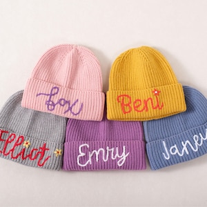 May include: A collection of colorful, knitted beanies. Each beanie is a different color, including pink, yellow, gray, purple, and blue. The names "Fox", "Beni", "Elliot", "Emry", and "Janeu" are embroidered on the front of each beanie in contrasting colors.