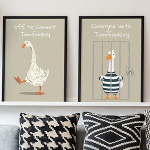 Może przedstawiać: Dwa oprawione plakaty z ilustracjami gęsi i tekstem. Jeden mówi "Off to Commit Tomfoolery", drugi "Charged with Tomfoolery". Plakaty znajdują się nad szarą sofą z wzorzystymi poduszkami.