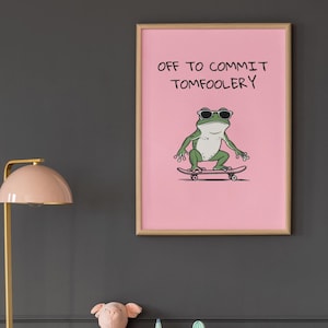 Może przedstawiać: Oprawiony różowy plakat z napisem "OFF TO COMMIT TOMFOOLERY" i kreskówkową żabą w okularach przeciwsłonecznych na deskorolce. Plakat znajduje się nad szarą komodą z elementami dekoracyjnymi.