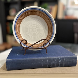 May include: A white ceramic bowl with brown and blue concentric rings, displayed on a decorative, wrought-iron stand. The bowl is resting on a vintage blue book. The bowl's design features a spiral pattern in the center.