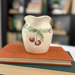 May include: A small, off-white ceramic vase with a wavy rim, adorned with a hand-painted cherry design. The cherries are a deep red, and the leaves are green. The vase is sitting on top of a stack of books.