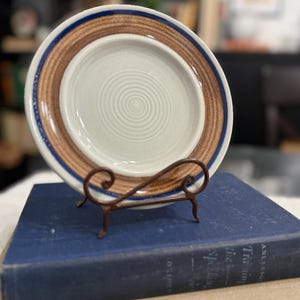 Op de afbeelding: Een vintage keramische plaat met een bruine en blauwe rand, rustend op een decoratieve metalen standaard. De plaat heeft een lichtgekleurd midden met een concentrisch cirkelontwerp. De plaat staat op een blauw boek.