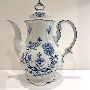 Könnte beinhalten: Weiße Porzellan-Kaffeekanne mit blauem Blumenmuster. Die Kanne hat einen gebogenen Ausguss, einen dekorativen Griff und einen Deckel mit Knauf. Das blaue Blumenmuster umfasst Blumen, Blätter und Ranken, was einen klassischen, eleganten Look erzeugt.