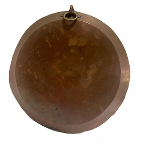 Może przedstawiać: Okrągły, miedziany metalowy talerz z lekko nieregularną krawędzią. Talerz ma matowy wygląd i mały pierścień u góry do zawieszenia. Powierzchnia wykazuje ślady zużycia i wieku.