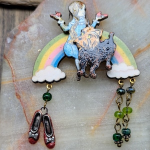 Peut inclure: Une broche arc-en-ciel colorée avec une fille et un chien sur le dessus. La fille porte une robe bleue et des chaussures rouges. Le chien est noir et brun. La broche a des perles vertes et dorées qui pendent.