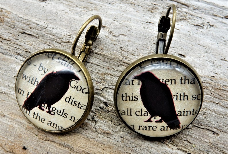 May include: A pair of bronze earrings with a round glass charm featuring a black silhouette of a raven on a vintage book page. The text on the page reads "God dista - by with mtangels n in the ans" and "at this all cla ven tha with so aint a rare an".