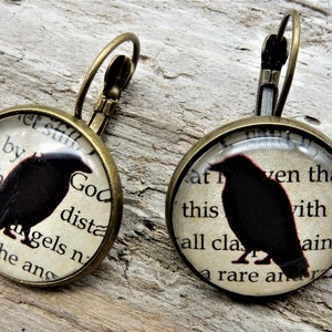 May include: A pair of bronze earrings with a round glass charm featuring a black silhouette of a raven on a vintage book page. The text on the page reads "God dista - by with mtangels n in the ans" and "at this all cla ven tha with so aint a rare an".