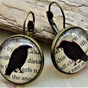 May include: A pair of bronze dangle earrings with a round glass charm featuring a black silhouette of a raven on a vintage book page background. The text on the page reads "et still s-by with God dista mt angels n om the ans" and "At ven tha this vith so all cla ain rare an".