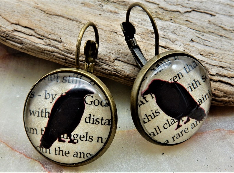 May include: A pair of bronze dangle earrings with a round glass charm featuring a black silhouette of a raven on a vintage book page background. The text on the page reads "et still s-by with God dista mt angels n om the ans" and "At ven tha this vith so all cla ain rare an".