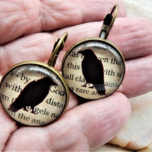 May include: A pair of bronze earrings with a black crow silhouette on a vintage book page background. The text on the page reads "God dista" and "a rare an".