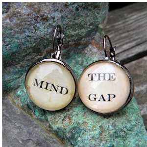 Puede incluir: Un par de pendientes de plata con abalorios redondos de cristal. Los abalorios son de color crema con texto negro. El pendiente izquierdo dice "MIND" y el pendiente derecho dice "THE GAP".