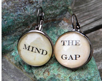 Puede incluir: Un par de pendientes de plata con abalorios redondos de cristal. Los abalorios son de color crema con texto negro. El pendiente izquierdo dice "MIND" y el pendiente derecho dice "THE GAP".