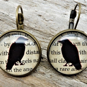 May include: A pair of bronze earrings with a glass cabochon featuring a black raven silhouette on a vintage book page background. The text on the page reads " - by God with dista om t angels na om the ans" and "at ven tha this with sc all cla ain te a rare an".